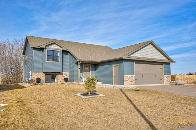 split level home featuring a front yard, roof with shingles, a garage, stone siding, and driveway