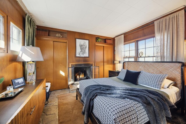 bedroom with stone finish floor, multiple windows, a warm lit fireplace, and wood walls