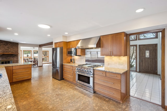 kitchen featuring wall chimney range hood, light stone countertops, decorative backsplash, appliances with stainless steel finishes, and brown cabinetry