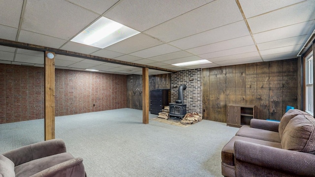 carpeted living area featuring a wood stove, brick wall, wood walls, and a drop ceiling