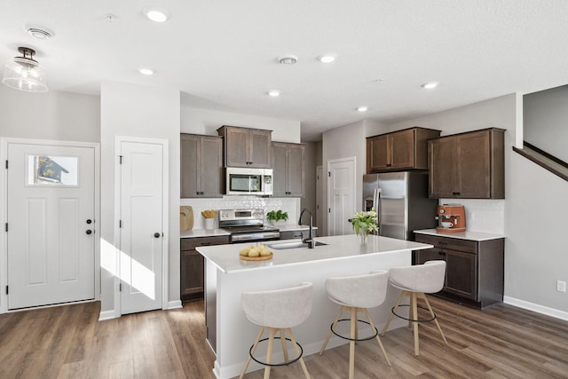 kitchen featuring a kitchen island with sink, a sink, wood finished floors, stainless steel appliances, and decorative backsplash