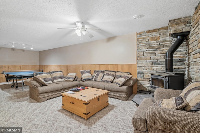 living room featuring a wainscoted wall, a textured ceiling, light colored carpet, ceiling fan, and a wood stove