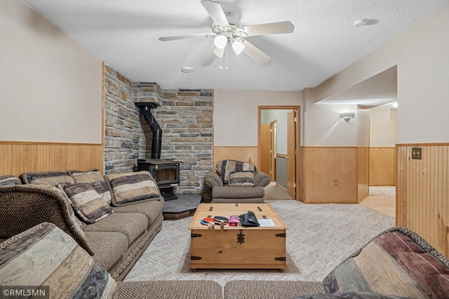 living room featuring a textured ceiling, wooden walls, wainscoting, ceiling fan, and a wood stove