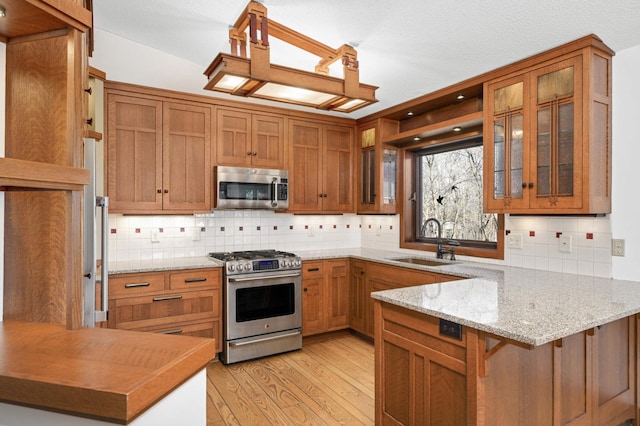 kitchen with light wood-style flooring, appliances with stainless steel finishes, a peninsula, brown cabinetry, and a sink