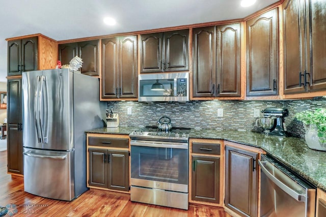 kitchen with light wood finished floors, tasteful backsplash, dark brown cabinets, dark stone countertops, and appliances with stainless steel finishes