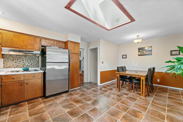 kitchen featuring under cabinet range hood, light countertops, decorative backsplash, freestanding refrigerator, and brown cabinetry