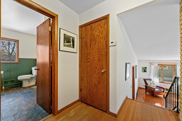 hallway with wood finished floors, a wainscoted wall, baseboards, a textured ceiling, and an upstairs landing