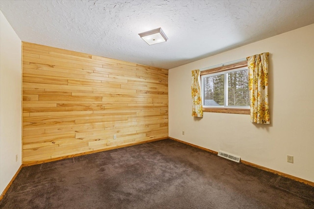 empty room featuring baseboards, visible vents, wood walls, a textured ceiling, and dark carpet