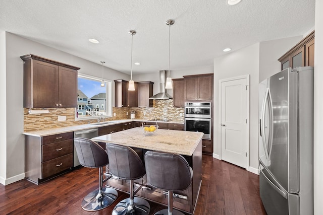 kitchen with wall chimney range hood, dark wood-style flooring, appliances with stainless steel finishes, and a kitchen island