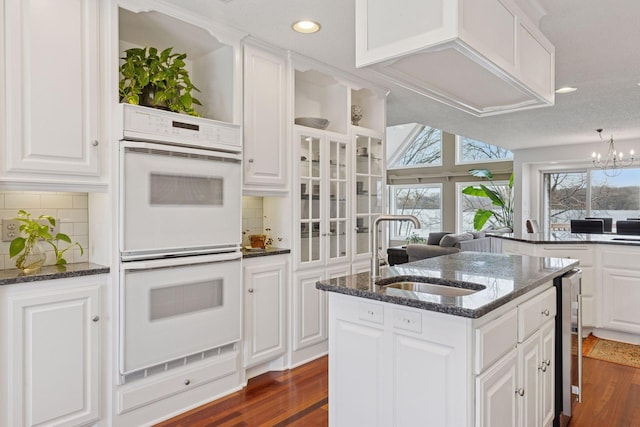 kitchen featuring an island with sink, a sink, backsplash, double oven, and dark wood-style flooring