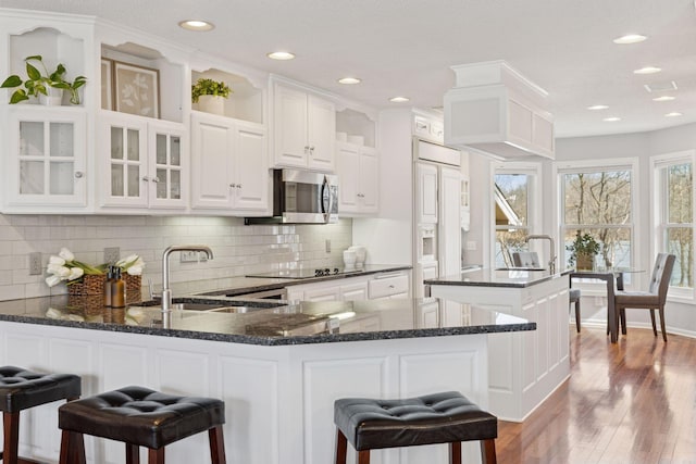 kitchen featuring stainless steel microwave, a peninsula, wood finished floors, white cabinetry, and a sink