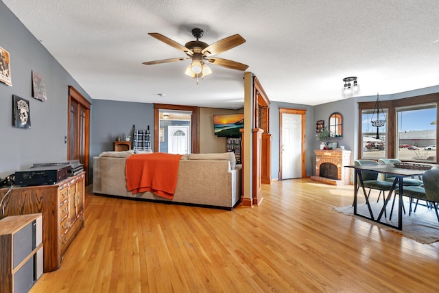 living area with a textured ceiling, light wood-style flooring, a brick fireplace, and a ceiling fan
