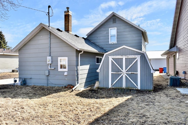 rear view of property featuring a shingled roof, a shed, central air condition unit, a chimney, and an outbuilding