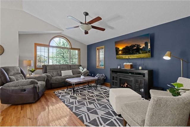 living room featuring a glass covered fireplace, hardwood / wood-style floors, a ceiling fan, and vaulted ceiling