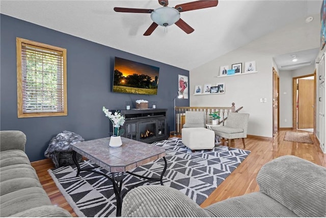 living room featuring a warm lit fireplace, wood finished floors, baseboards, ceiling fan, and vaulted ceiling