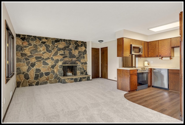 kitchen featuring brown cabinets, stainless steel appliances, a stone fireplace, light colored carpet, and light countertops