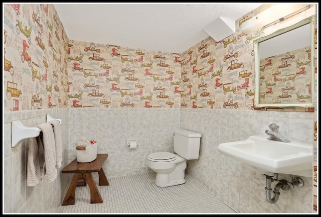bathroom featuring toilet, wallpapered walls, tile walls, a textured ceiling, and wainscoting