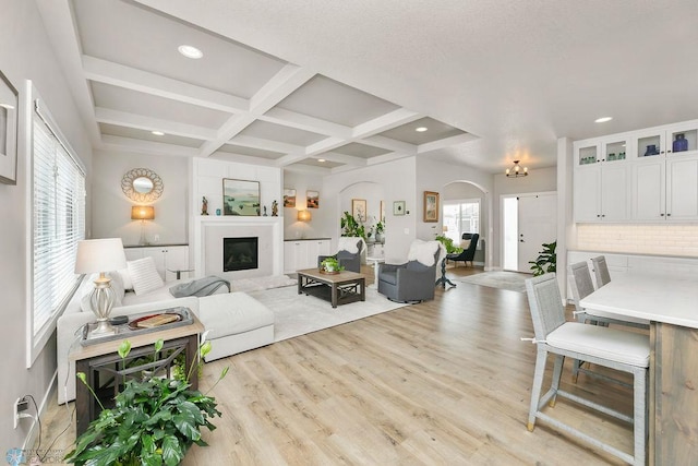 living area with coffered ceiling, a fireplace with raised hearth, arched walkways, beamed ceiling, and light wood-type flooring