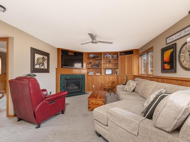 carpeted living area featuring built in shelves, a fireplace with flush hearth, a ceiling fan, wood walls, and wainscoting