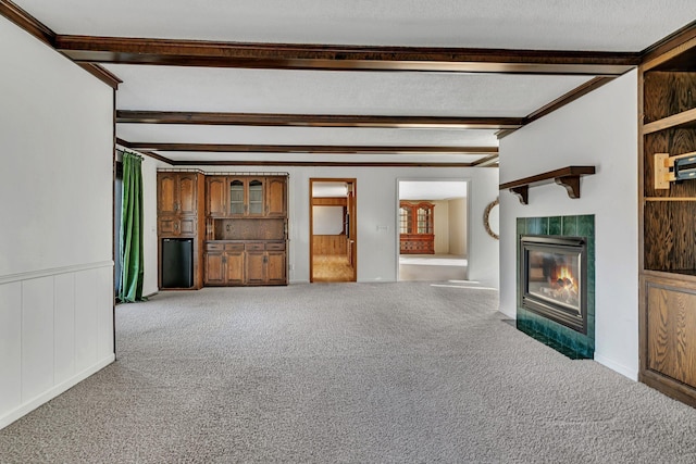 unfurnished living room with beamed ceiling, a tiled fireplace, a textured ceiling, carpet, and wainscoting