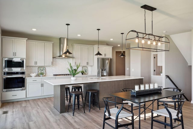 kitchen with a center island with sink, stainless steel appliances, wall chimney exhaust hood, light countertops, and decorative backsplash