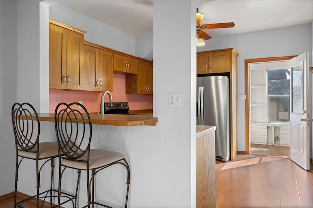 kitchen with a ceiling fan, wood finished floors, a peninsula, stainless steel fridge with ice dispenser, and a kitchen bar