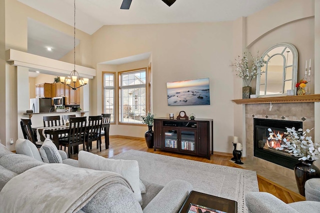 living room featuring baseboards, a tiled fireplace, light wood-type flooring, ceiling fan with notable chandelier, and high vaulted ceiling