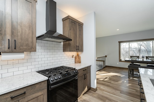 kitchen featuring black range with gas cooktop, wall chimney range hood, light wood-type flooring, decorative backsplash, and baseboards