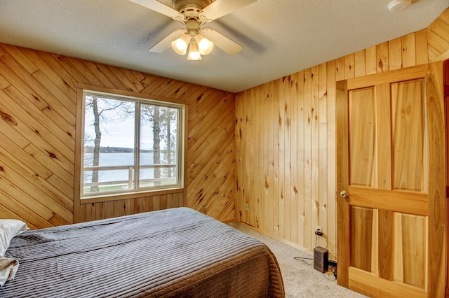 carpeted bedroom with a textured ceiling, a water view, wooden walls, and ceiling fan