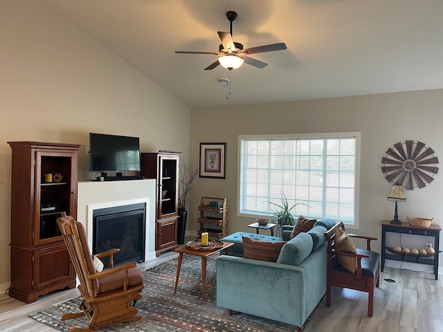 living area featuring a glass covered fireplace, vaulted ceiling, light wood finished floors, and ceiling fan