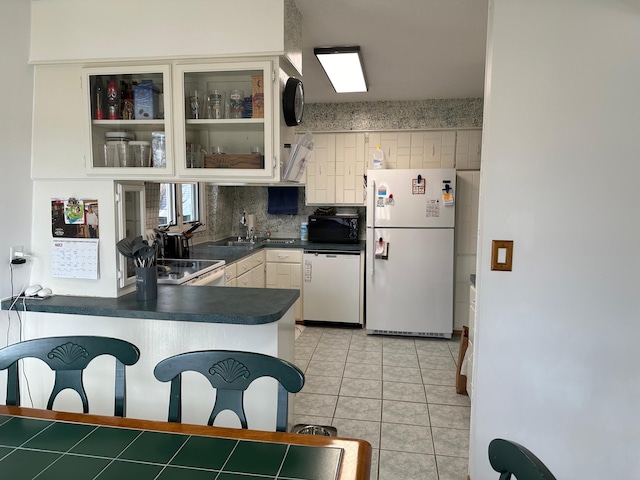 kitchen with decorative backsplash, a peninsula, dark countertops, white appliances, and glass insert cabinets