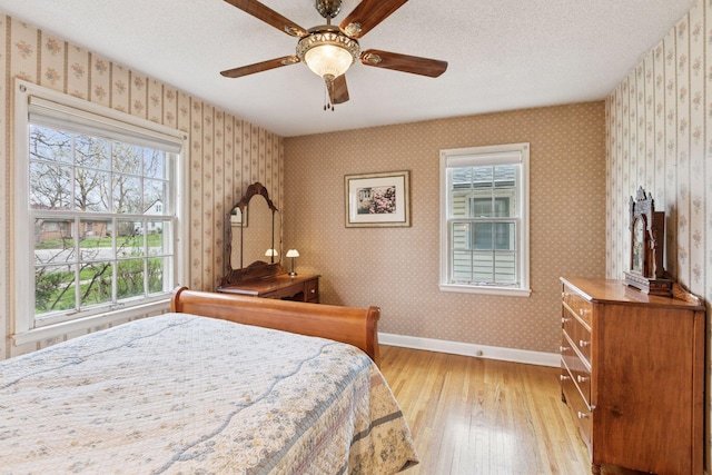 bedroom featuring a textured ceiling, light wood-type flooring, ceiling fan, baseboards, and wallpapered walls