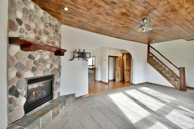 unfurnished living room featuring carpet, arched walkways, a ceiling fan, a stone fireplace, and wooden ceiling