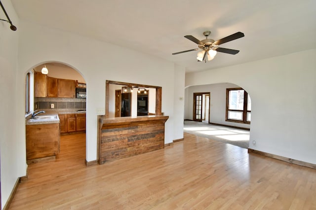 unfurnished living room with baseboards, arched walkways, a sink, ceiling fan, and light wood-style floors