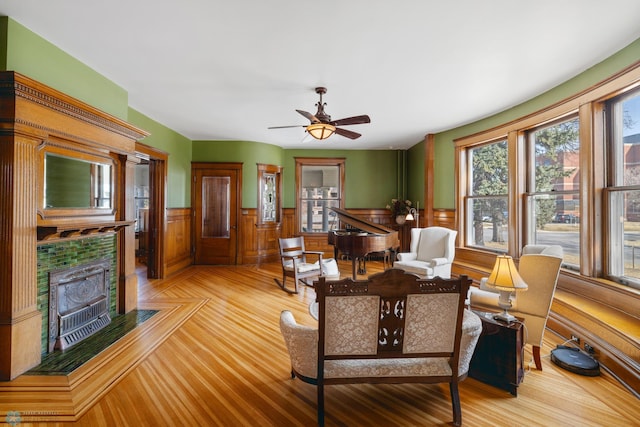 living area featuring a tiled fireplace, a wainscoted wall, and ceiling fan