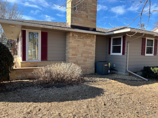 exterior space with central air condition unit, a chimney, a shingled roof, and stone siding