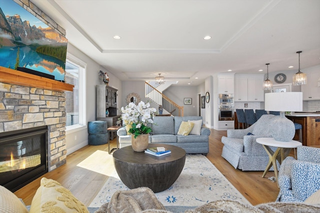 living area featuring a tray ceiling, a stone fireplace, light wood finished floors, and stairway
