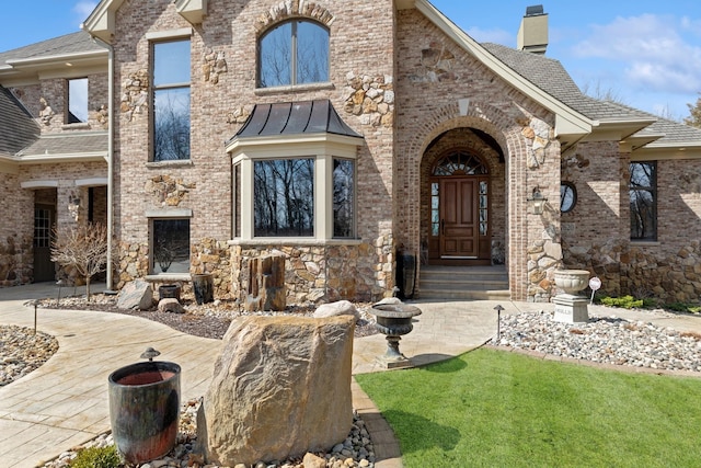 doorway to property with a lawn, brick siding, a chimney, and roof with shingles