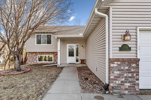 doorway to property featuring a shingled roof, an attached garage, and brick siding