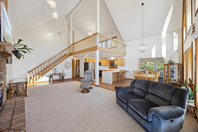living area with high vaulted ceiling, stairway, light wood-style floors, a chandelier, and a brick fireplace