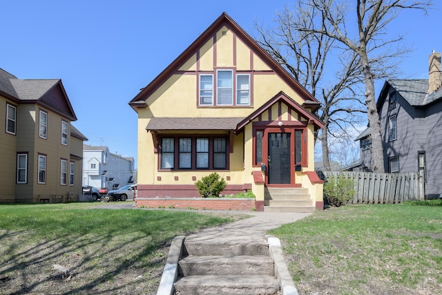 english style home featuring fence, stucco siding, entry steps, and a front lawn