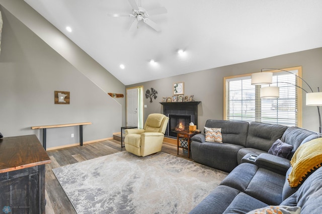 living area featuring ceiling fan, vaulted ceiling, wood finished floors, baseboards, and a warm lit fireplace