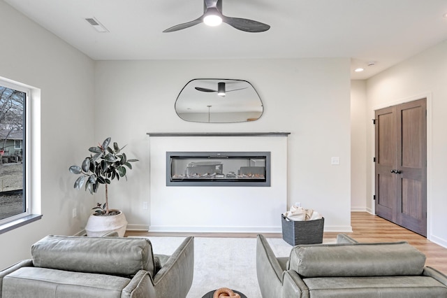 living room with a ceiling fan, a glass covered fireplace, light wood-type flooring, baseboards, and visible vents