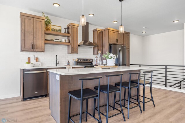kitchen with wall chimney range hood, appliances with stainless steel finishes, a center island, a sink, and light wood finished floors