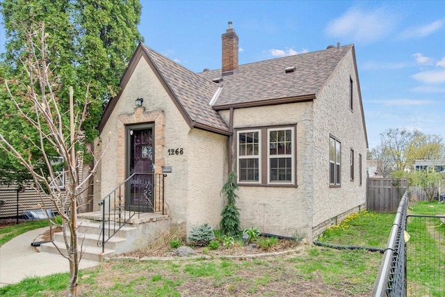 english style home with fence, stucco siding, a chimney, and roof with shingles