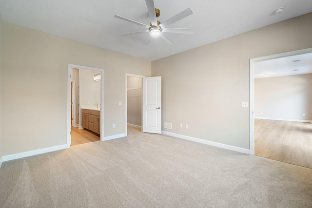 unfurnished bedroom featuring a walk in closet, baseboards, ensuite bath, ceiling fan, and light carpet