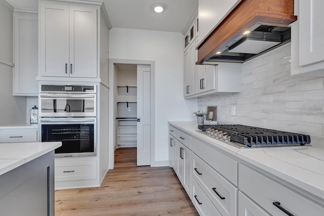kitchen with custom range hood, tasteful backsplash, light wood finished floors, appliances with stainless steel finishes, and light stone countertops