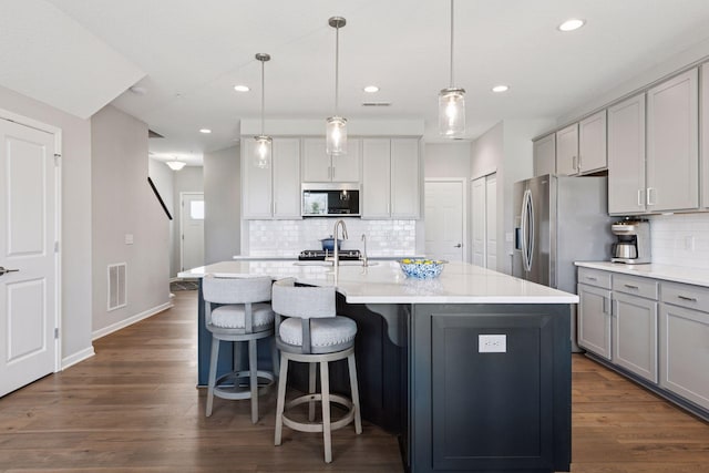 kitchen with a kitchen island with sink, stainless steel microwave, decorative backsplash, gray cabinetry, and dark wood-type flooring