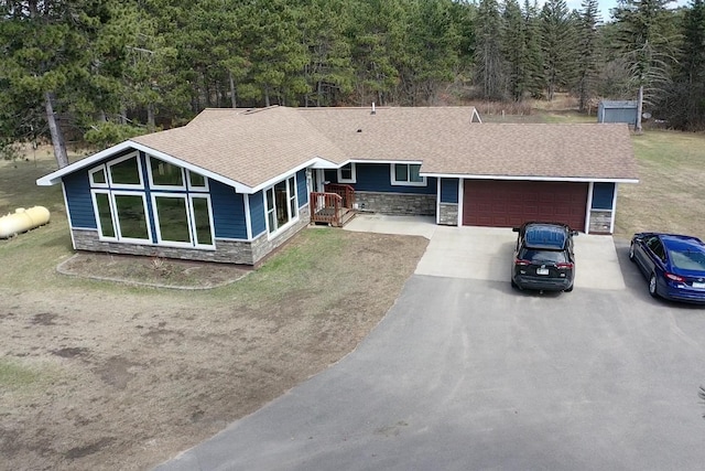 view of front facade featuring a garage, driveway, stone siding, a view of trees, and roof with shingles