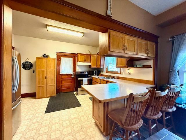 kitchen with light countertops, a peninsula, appliances with stainless steel finishes, under cabinet range hood, and brown cabinetry